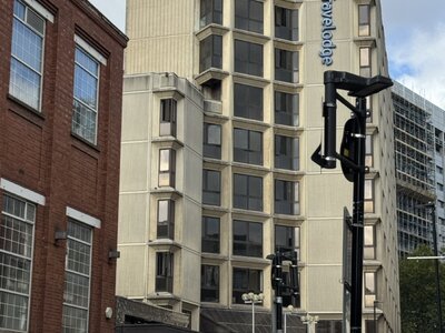 Tall beige Travelodge hotel building with vertical signage, white delivery van parked in front, red brick Victorian building to left, black street lamp in foreground, pedestrians on pavement