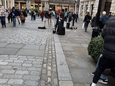 Cobblestone plaza with street musician equipment on ground, crowd of tourists watching, cream-colored classical building facades, ornate lamp post, Covent Garden piazza