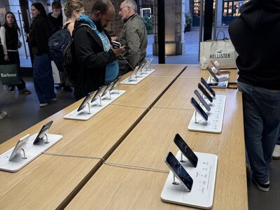 Apple Store interior with wooden display tables, rows of iPhones and iPads on white stands, customers browsing, large windows showing London architecture outside