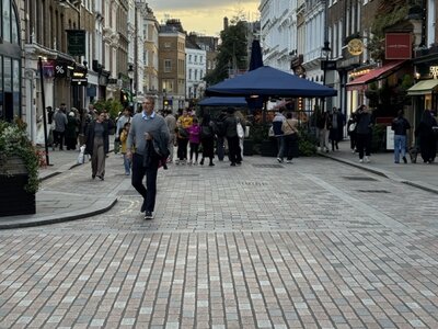 Cobblestone pedestrian street at dusk, blue event tent in center, Victorian and Georgian buildings on both sides, people walking, dramatic cloudy sky