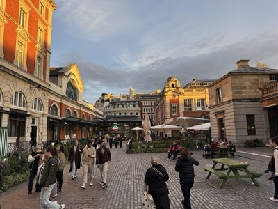 Covent Garden piazza at sunset, cobblestone square, red London Underground roundel sign, outdoor restaurant seating, white umbrellas, classical facades, warm light