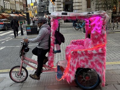 Pink pedicab rickshaw covered in fluffy faux fur with colorful LED light strips, parked on street, traffic and pedestrians in background