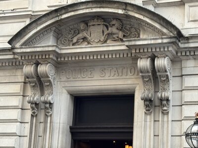 Stone archway entrance with royal coat of arms carved in relief, ionic column capitals, ornamental moldings, POLICE STATION inscription above arch