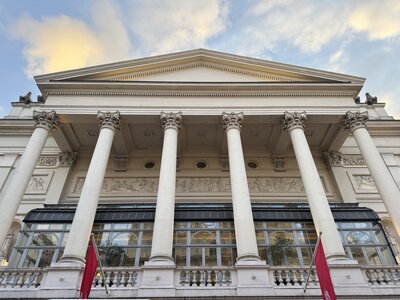 Royal Opera House with neoclassical facade, six towering Corinthian columns, triangular pediment with sculptural reliefs, red banners hanging from columns, tourists on forecourt