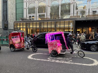 Two pink pedicab rickshaws with LED lights and faux fur coverings parked in front of Royal Opera House ornate glass and iron facade, Covent Garden street scene