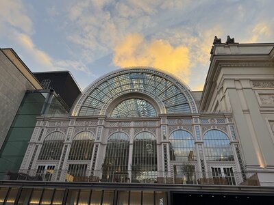 Royal Opera House Covent Garden with ornate Victorian glass and iron facade, large semi-circular arched windows with decorative ironwork, dramatic sunset clouds with golden light in background