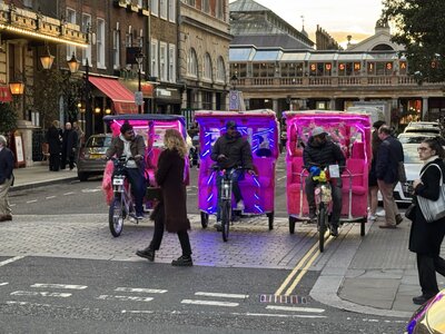 Three LED-lit pedicab rickshaws with pink and purple neon lights at dusk, woman in dark coat crossing street, Sushi SA restaurant background, traffic