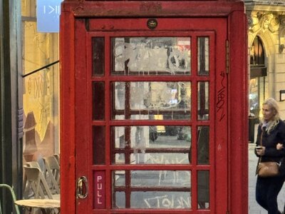 Classic British red K6 telephone box on pavement, cafe seating in middle distance, illuminated commercial building facades, daytime urban London scene