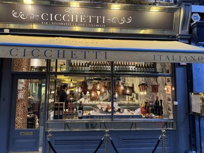 Cicchetti Italian restaurant storefront at dusk, elegant blue and cream facade, windows displaying wine bottles, hanging prosciutto, pendant lights, BAR RISTORANTE CAFFE signage