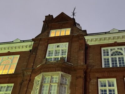 Victorian red brick building at dusk, warm lights glowing in windows, distinctive tower spire silhouetted against evening sky, ornate 19th century architecture
