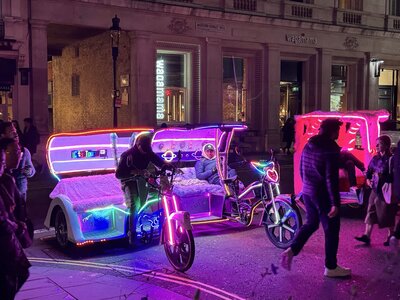 LED-illuminated pedicab rickshaws at night with multicolor neon light strips, passengers seated, Wagamama Asian restaurant signage glowing in background