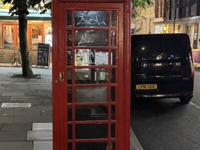 Classic British red K6 telephone box in evening with graffiti on glass panels, black van parked behind, tree and illuminated shop windows