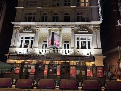 Duke of York's Theatre illuminated at night, grand neoclassical white facade, large STEREOPHONIC marquee sign, golden columns, ornate balustrade, purple seating, West End