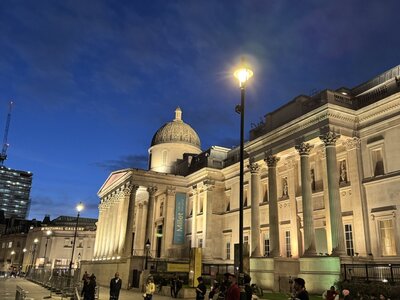 National Gallery museum at Trafalgar Square illuminated at dusk, distinctive dome, neoclassical facade, towering Corinthian columns, yellow Miller banners, people on forecourt