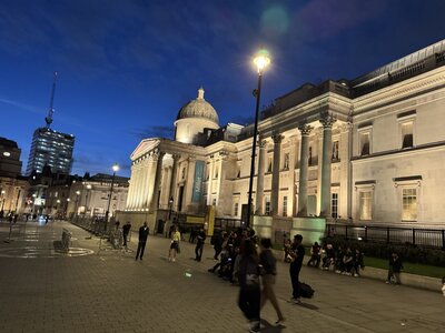 National Gallery at Trafalgar Square at dusk, neoclassical facade, prominent dome, Corinthian columns, yellow Miller exhibition banners, pedestrians and photographers, blue evening sky