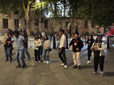 Public square at night with young people performing coordinated group dance on cobblestone plaza, audience watching, neoclassical government building, ornate lamps, trees