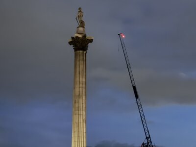 Nelson's Column at Trafalgar Square at dusk, tall fluted Corinthian column against dramatic cloudy sky, illuminated historic buildings, construction crane, monument