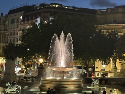 Illuminated fountain at Trafalgar Square at night with water jets spraying, trees and historic buildings in background, dramatic evening atmosphere, people gathering around monument, warm street lighting