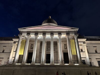 National Gallery museum at night with dramatic illumination, neoclassical facade, distinctive dome, towering Corinthian columns, yellow banners, people on Trafalgar Square forecourt
