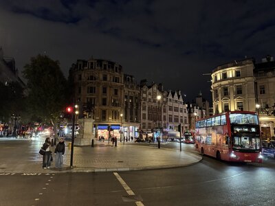 London street scene at night with illuminated historic buildings, red double-decker bus passing through intersection, street lamps glowing, pedestrians and traffic