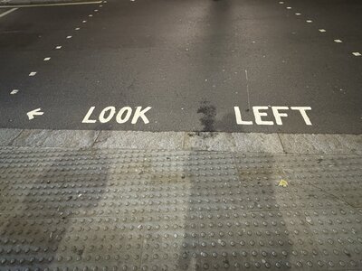 Large white LOOK LEFT road surface marking with directional arrow painted on dark asphalt, grey tactile paving for visually impaired users along curb