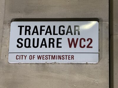 White street sign with black lettering TRAFALGAR SQUARE WC2 mounted on weathered stone wall, red text CITY OF WESTMINSTER designation below