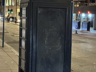 Black modernized telephone kiosk with WiFi here sign illuminated on top, royal crown emblem, standing on pavement at night, illuminated neoclassical building background