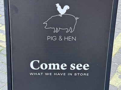 Black A-frame sidewalk sign for Pig & Hen jewelry brand with white logo of chicken standing on pig, text Come see what we have in store, Handmade Jewelry from Amsterdam, street advertising board