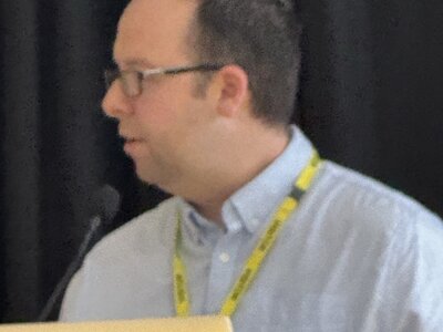 TYPO3 conference attendee wearing glasses and light shirt with yellow lanyard working on MacBook laptop, focused on screen, dark background curtain, technical session