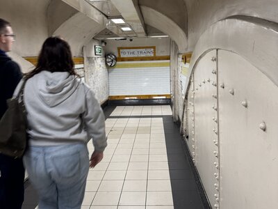 London Underground tunnel passage with curved arched ceiling, white tiled walls, pedestrian walking towards closed shuttered gate marked TO THE TRAINS, floor clock, yellow accent stripes