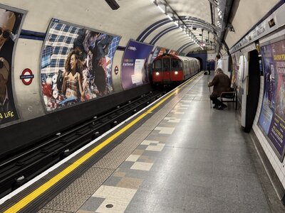 London Underground platform with red tube train arriving at station, curved tunnel ceiling, colorful advertising posters on tiled walls, passenger seated on bench waiting