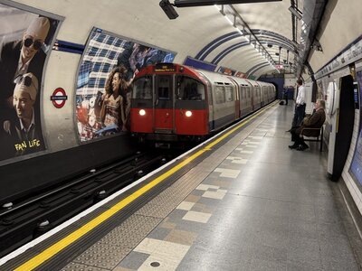 London Underground station platform with red and grey tube trains stopped, curved tunnel ceiling design, advertising posters lining walls, passengers waiting for trains