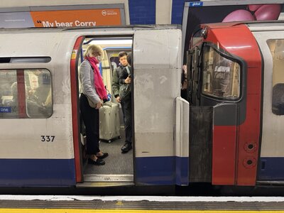 Female passenger with rolling suitcase luggage entering London Underground train through open doors, tube platform with advertising billboards on curved walls, commuter scene