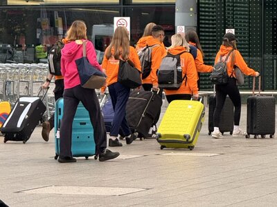 International tour group wearing matching bright orange high-visibility jackets carrying colorful wheeled luggage suitcases, assembled outside airport terminal building, organized group travel